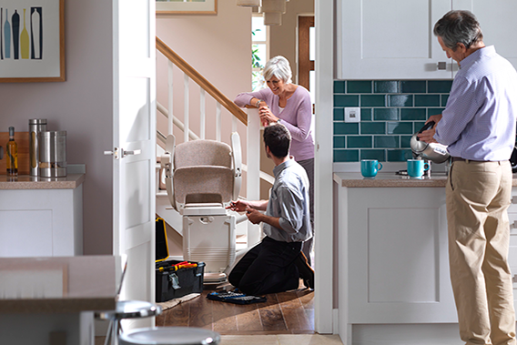 Senior woman smiling, watching a Stannah stairlift being serviced. His husband is nearby, in the kitchen, serving coffee.