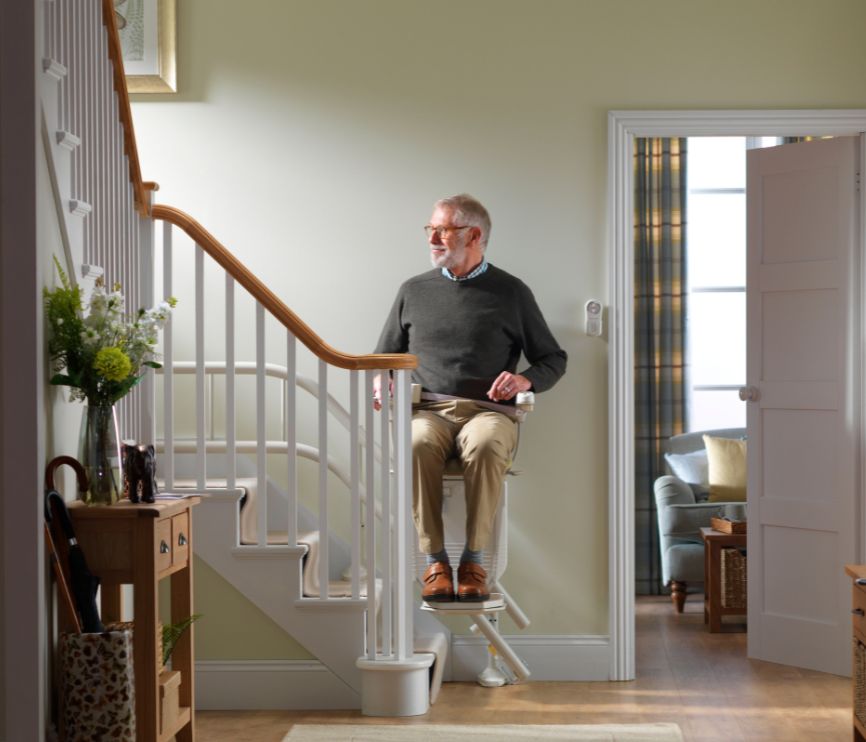 Older man seated on a Stannah stairlift at the foot of a curved staircase in a bright hallway, preparing to travel upstairs.