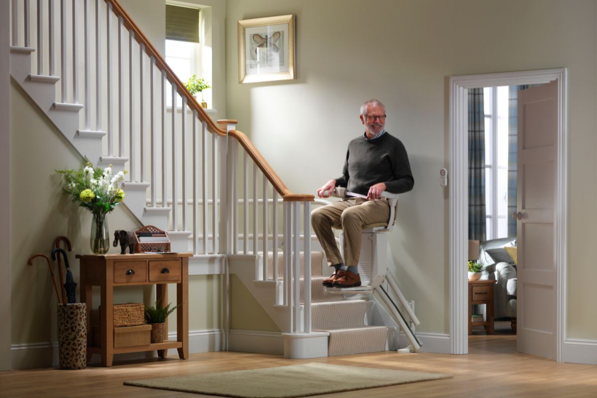Older man riding a Stannah stairlift around a curved staircase in a bright hallway, passing a small landing with white spindles and a wooden handrail.