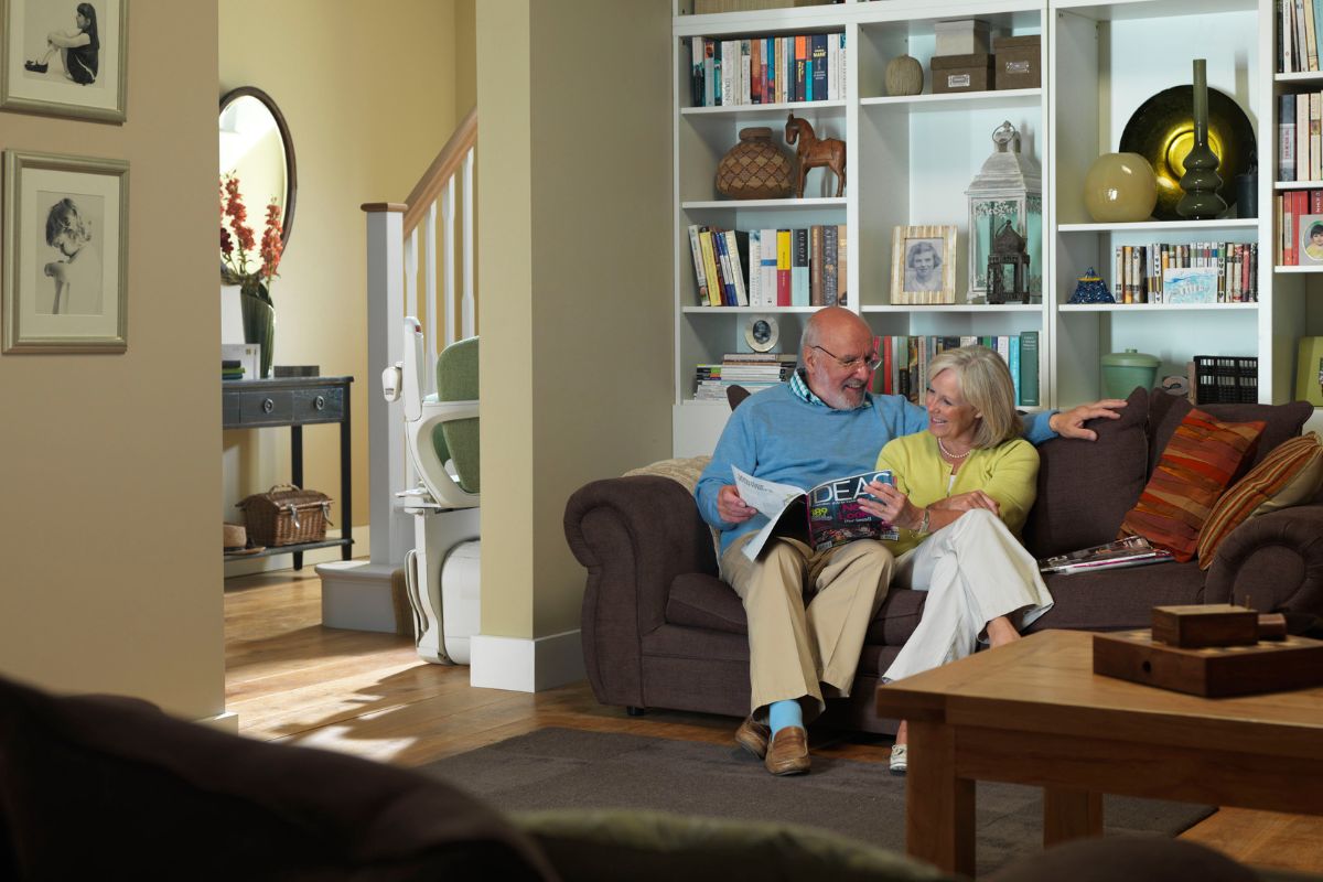 Older couple reading on a sofa in a cozy living room, with a Stannah stairlift (green seat) visible by the hallway staircase in the background.