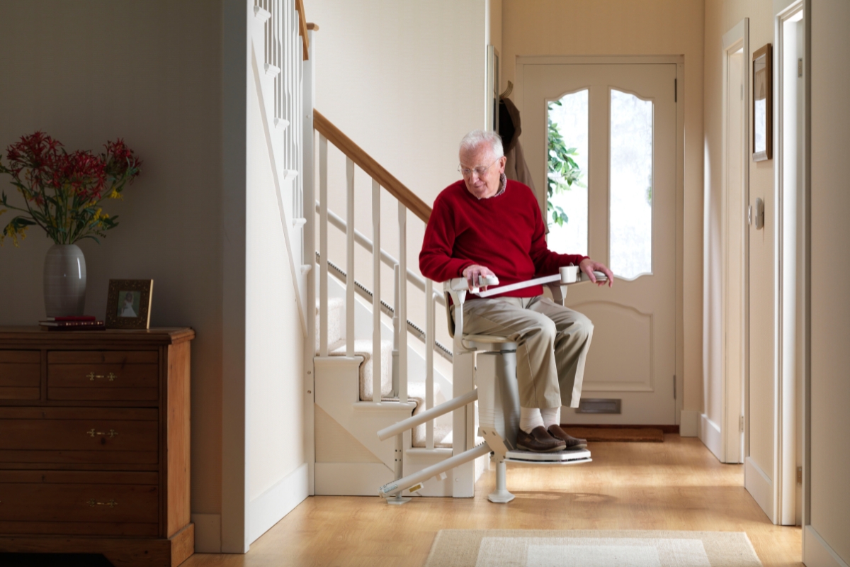 Older man riding a straight stairlift beside the front door in a bright hallway.